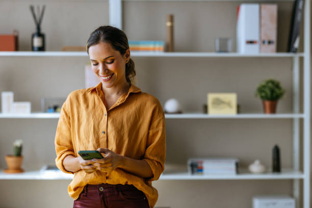 A smiling Caucasian entrepreneur texting on her smartphone while standing indoors.